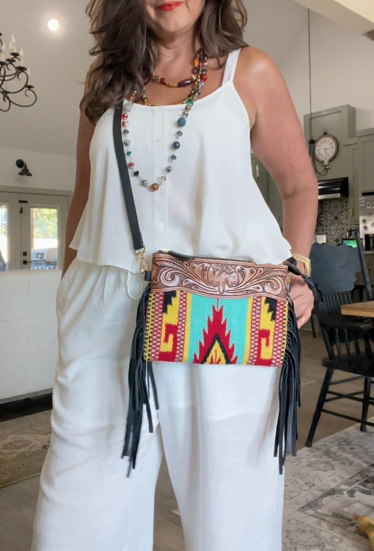 A woman wearing a white sleeveless top and white pants carries a colorful embroidered leather crossbody purse with black fringe. The bag features a turquoise, red, yellow, and brown geometric pattern with western-style embroidery on the strap.